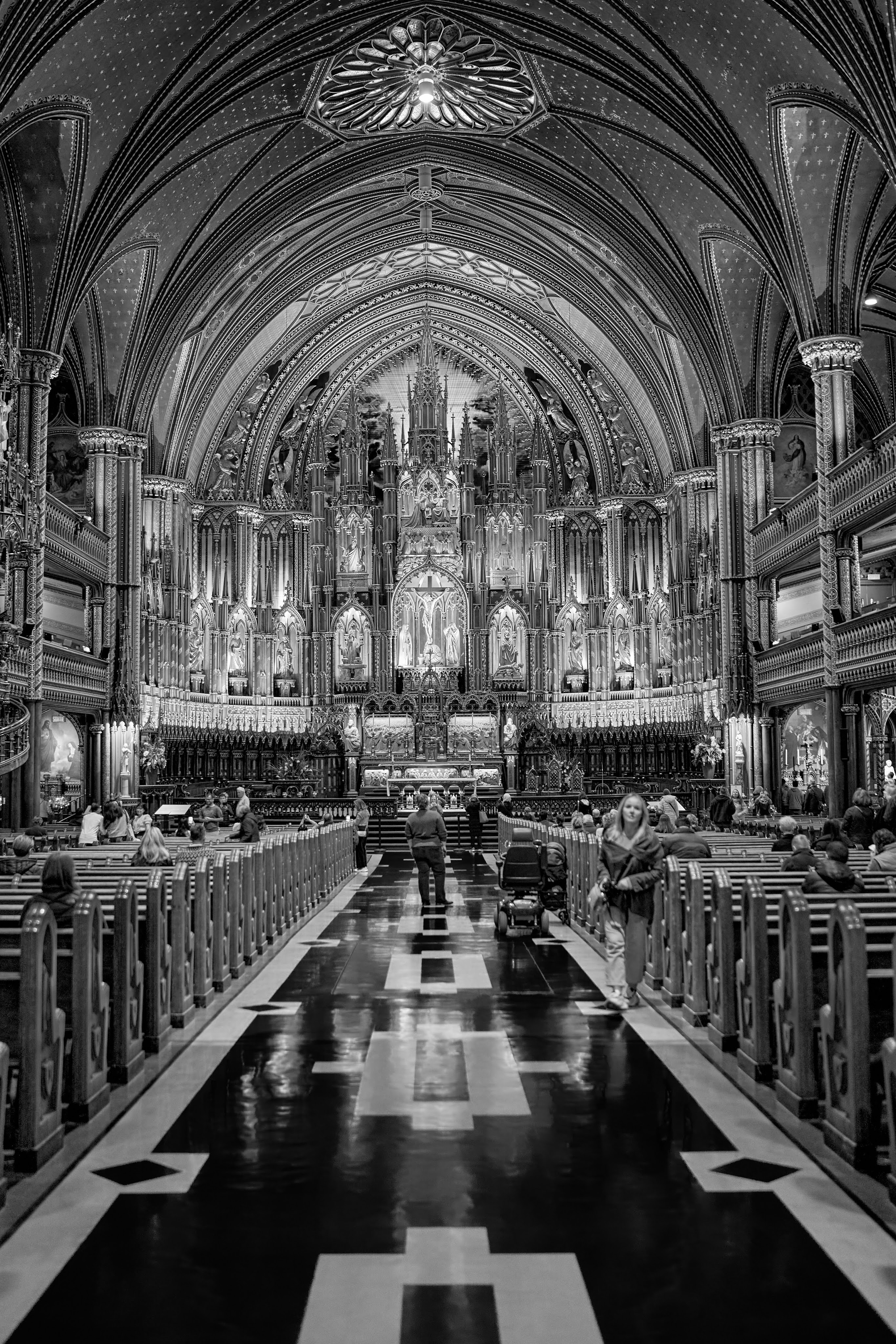 This is an elegant black-and-white photograph capturing the interior of the Notre-Dame Basilica of Montreal. The image showcases the intricate Gothic Revival architecture, ornate altar details, and soaring arches, with the delicate interplay of light and shadow adding depth. Visitors in the foreground provide a sense of scale and add a quiet, contemplative atmosphere to the grandeur of the basilica.